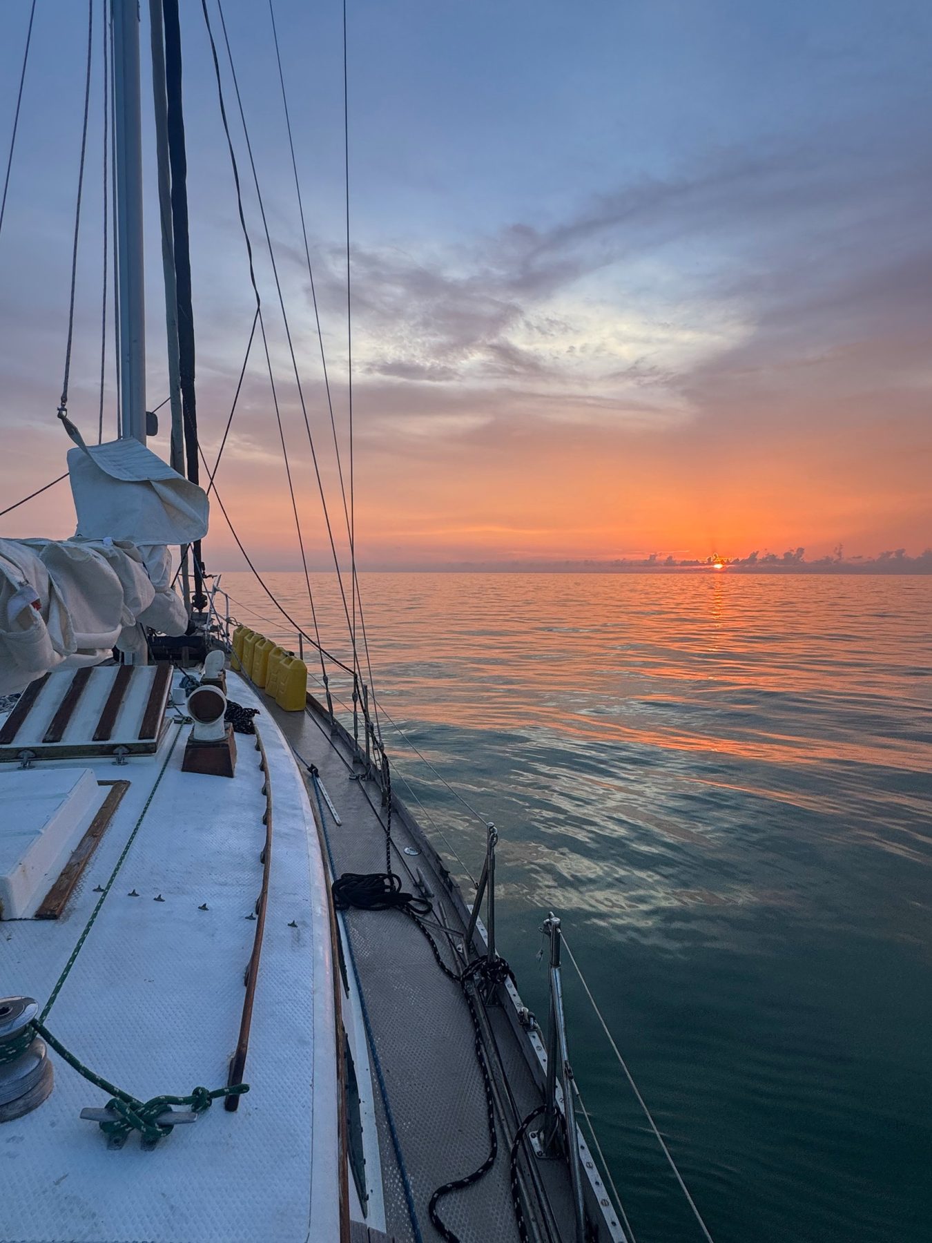 Orange sunset over calm water from Yemaya's foredeck, jerry cans lashed along the rail